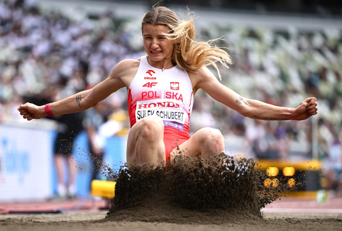Zawodniczka reprezentująca Polskę w trakcie lądowania podczas skoku w dal na stadionie lekkoatletycznym, piasek unosi się w górę, w tle rozmyte trybuny z kibicami.