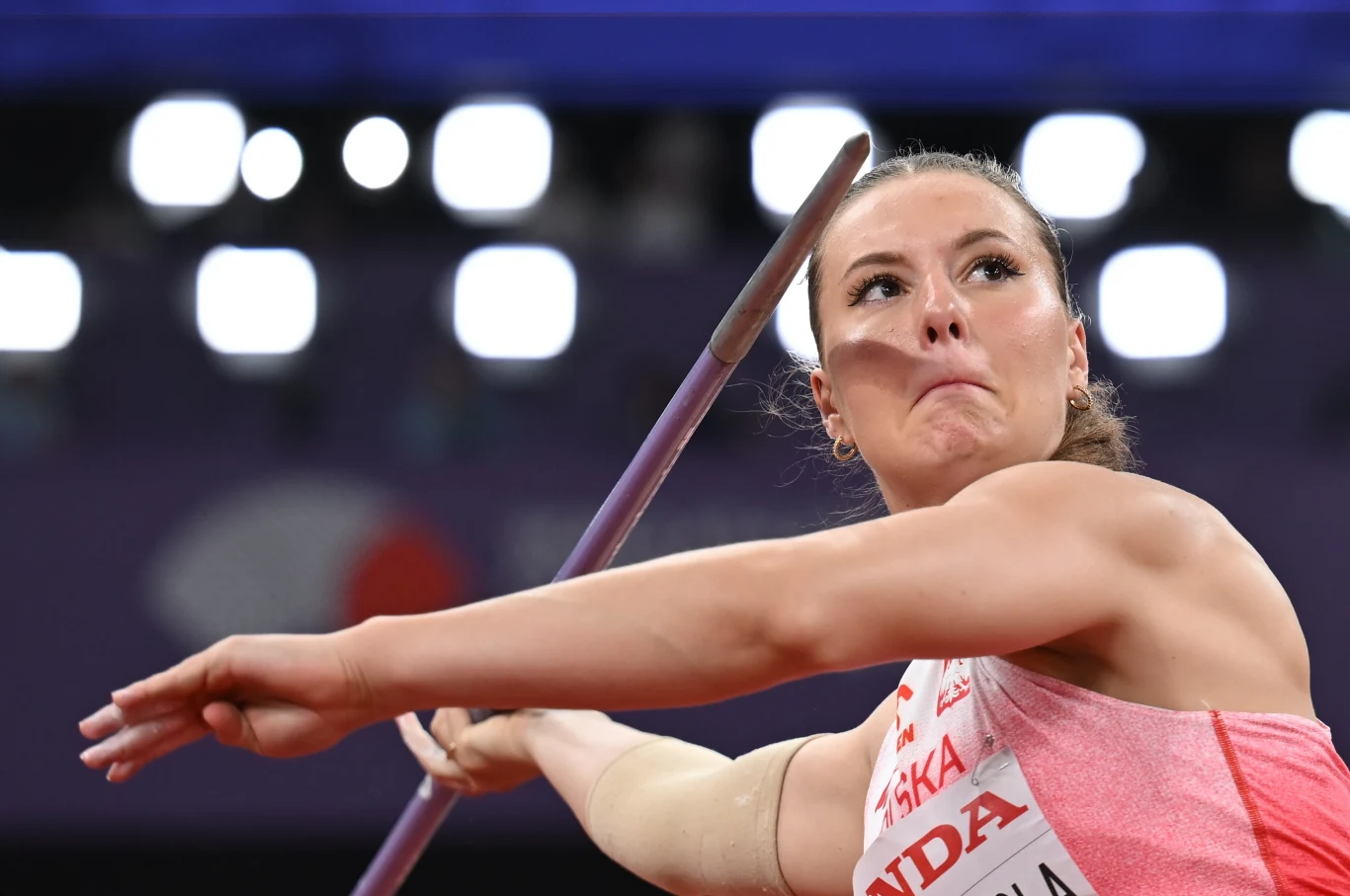 Zbliżenie na lekkoatletkę w trakcie rzutu oszczepem, skupienie i determinacja malujące się na twarzy, dynamiczna poza sportowa, wyraźnie widoczny oszczep, intensywne światło w tle.