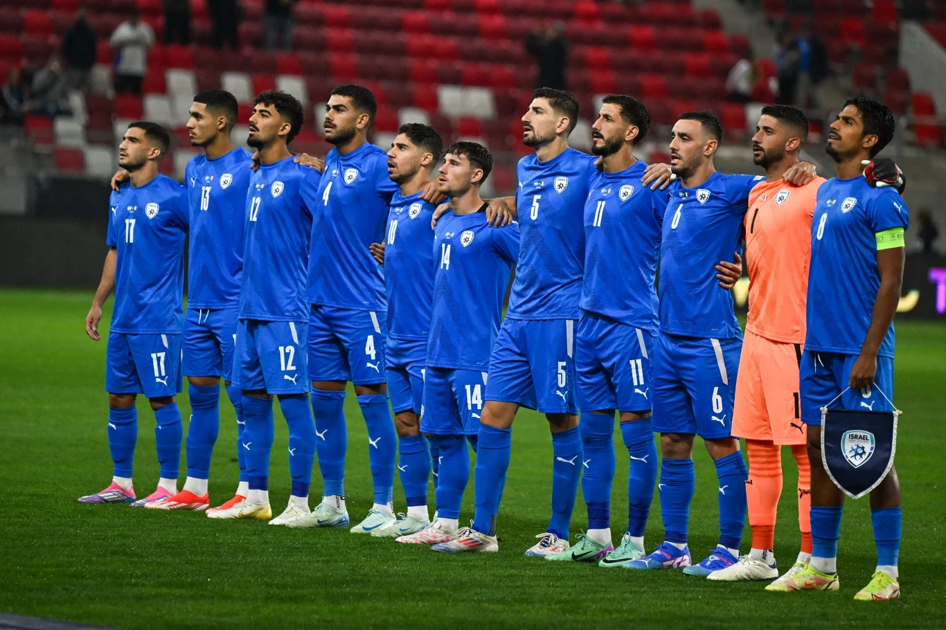 Piłkarze drużyny narodowej w niebieskich strojach stoją w szeregu, przygotowani do rozpoczęcia meczu na stadionie piłkarskim, z pustymi czerwonymi i szarymi trybunami w tle.