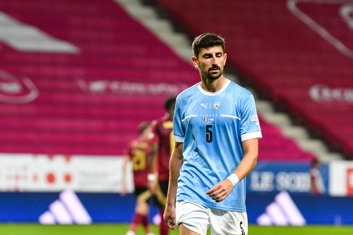 Piłkarz w jasnoniebieskiej koszulce z numerem 5 na przodzie, stojący na boisku podczas meczu. W tle rozmazana sylwetka innych zawodników i czerwone trybuny stadionu.