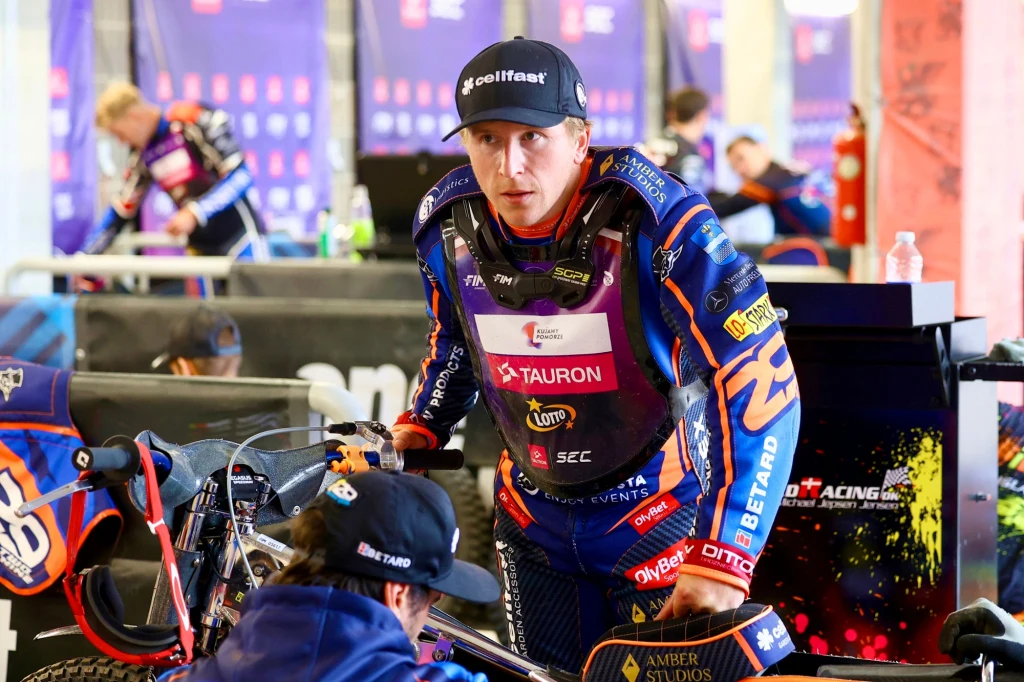 Zawodnik żużlowy w profesjonalnym stroju sportowym stoi obok swojego motocykla w parku maszyn, koncentrując się przed zawodami. W tle znajdują się inni członkowie zespołu oraz elementy wyposażenia technicznego.