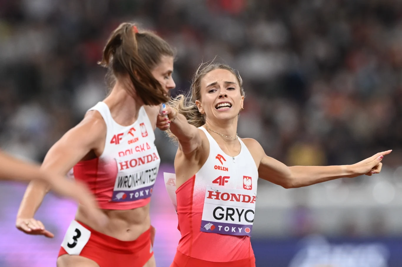 Dwie polskie lekkoatletki w trakcie wymiany pałeczki podczas biegu sztafetowego na stadionie, obie skoncentrowane, w strojach sportowych z widocznymi nazwiskami oraz barwami narodowymi.