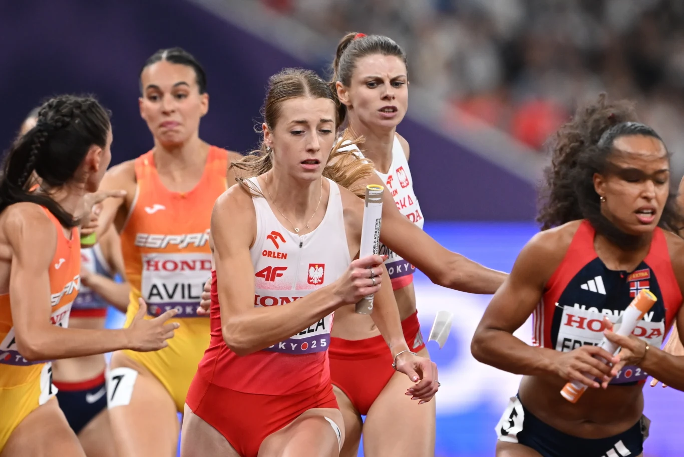 Zawodniczki biegnące w sztafecie lekkoatletycznej podczas zawodów, wyraziste emocje na twarzach i przekazywanie pałeczki między biegaczkami w otoczeniu stadionu sportowego.