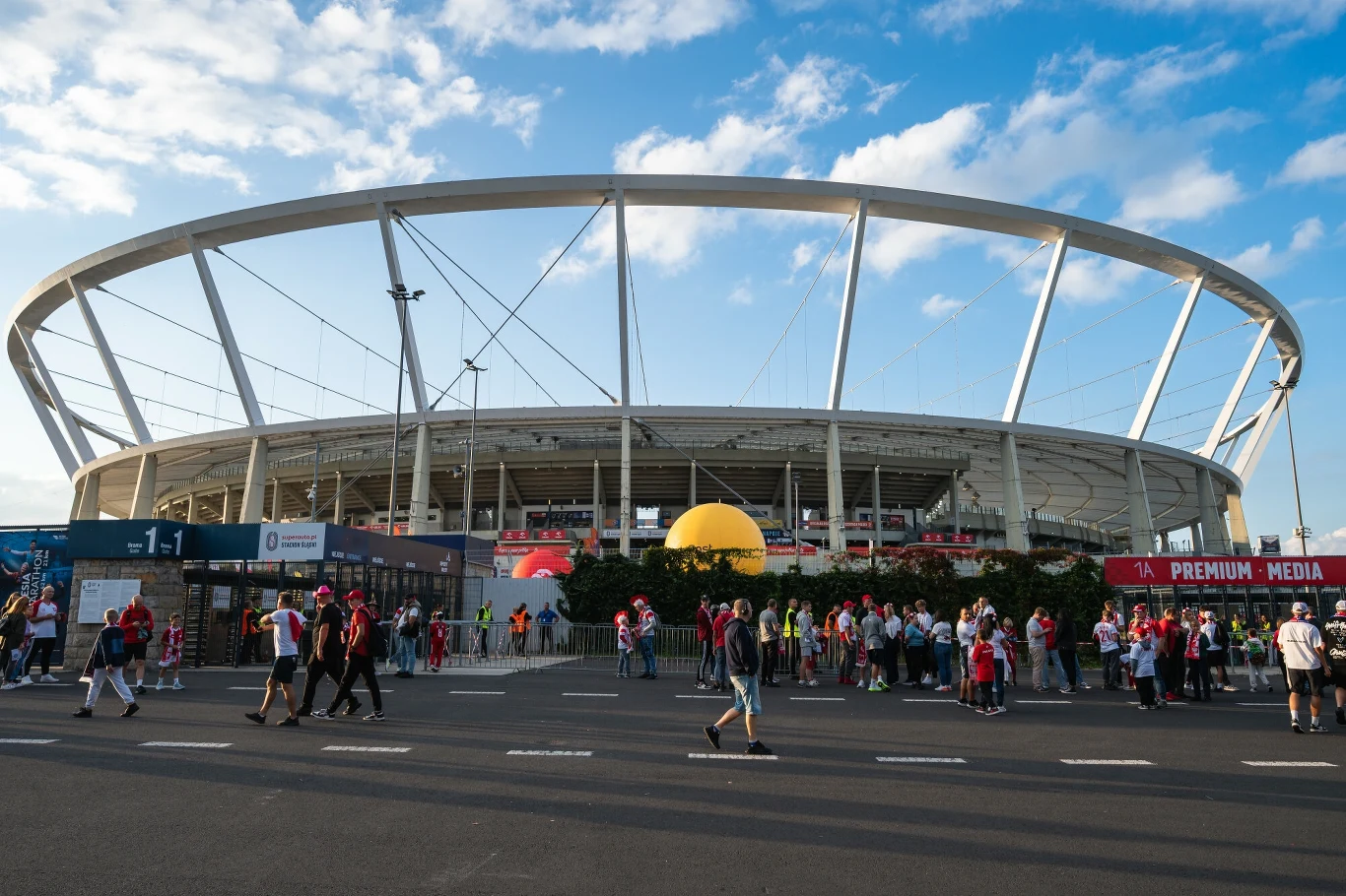 Stadion Śląski w Chorzowie Tłum ludzi zgromadzony przed nowoczesnym stadionem, niektórzy ubrani w kibicowskie barwy, na tle jasnego nieba z drobnymi chmurami.