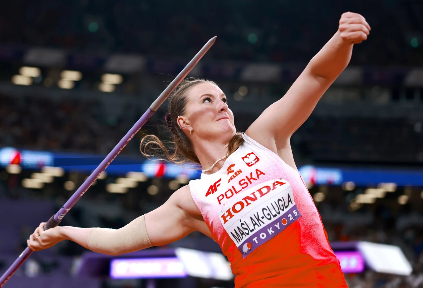 Lekkoatletka w biało-czerwonym stroju reprezentującym Polskę wykonuje rzut oszczepem na stadionie podczas zawodów, w tle rozmyta widownia i elementy infrastruktury sportowej.