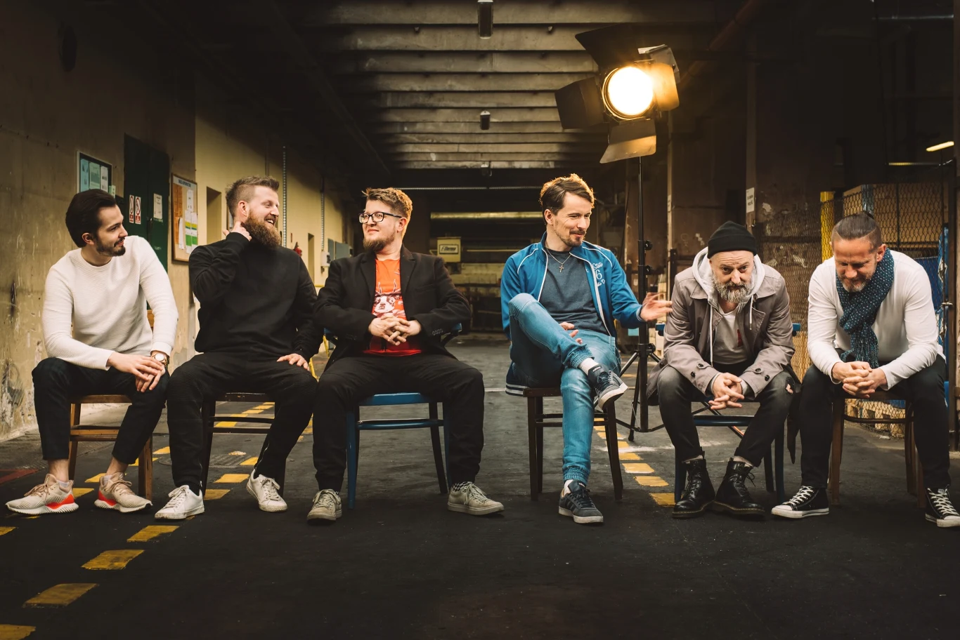 Akurat zagra koncert w Warszawie Grupa sześciu uśmiechniętych mężczyzn siedzących na krzesłach w rzędzie w industrialnym wnętrzu, nieformalna atmosfera, ciepłe oświetlenie tworzy przyjazny nastrój.