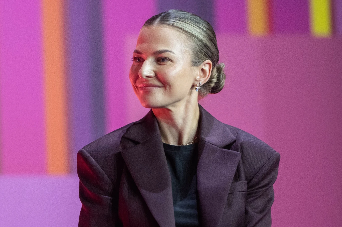 Anna Lewandowska Kobieta z blond włosami upiętymi w elegancki kok, ubrana w ciemny żakiet i czarną bluzkę, siedzi na tle różowych ścian z pionowymi, kolorowymi akcentami; uśmiecha się, patrząc lekko w bok.
