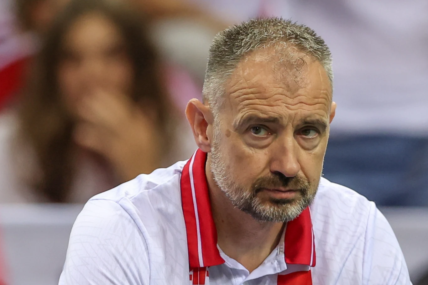 Mężczyzna w średnim wieku z siwiejącymi włosami i brodą ubrany w białą koszulkę z czerwonym akcentem na kołnierzu siedzi na trybunach, w tle widoczna rozmyta postać kobiety.