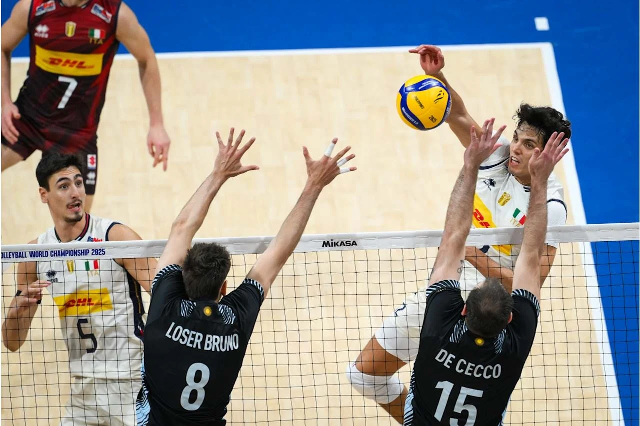Emocjonujący moment meczu siatkówki, jeden z zawodników atakuje piłkę nad siatką, podczas gdy dwóch rywali próbuje ją zablokować; widoczne są sportowe stroje i intensywna rywalizacja.