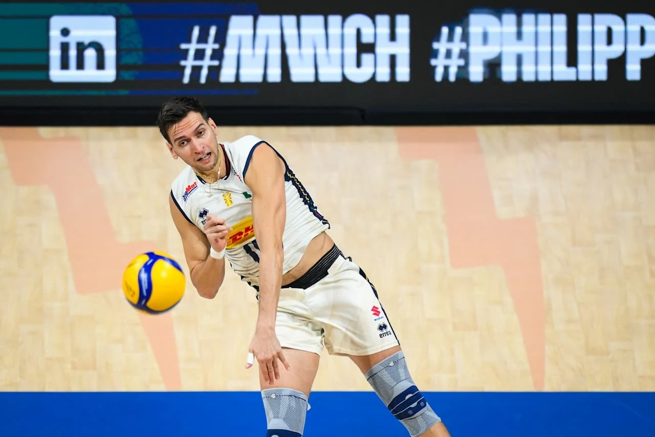 Siatkarz w stroju sportowym odbijający piłkę podczas meczu halowego, dynamiczna pozycja oraz wyraźne skupienie na grze; na tle widoczne oznaczenia i hashtagi na bandach reklamowych.