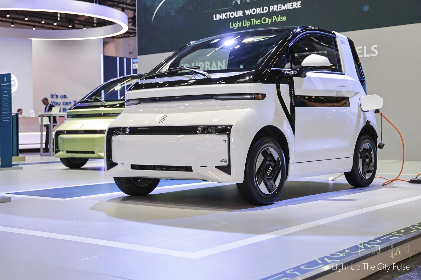 Nowoczesny, kompaktowy samochód elektryczny w białym kolorze zaprezentowany na targach motoryzacyjnych, widoczny kabel przyłączony do ładowania pojazdu, w tle inne auta elektryczne i elementy stoiska wystawienniczego.