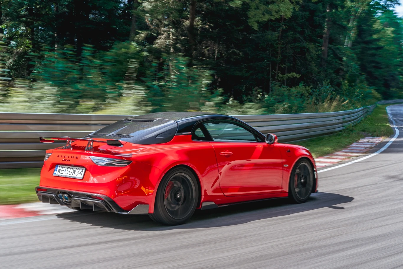 Alpine A110 R zwraca uwagę karbonowym pakietem aerodynamicznym w tym spojlerem na tylnej klapie Czerwony samochód sportowy poruszający się z dużą prędkością po torze wyścigowym otoczonym barierkami i zielenią