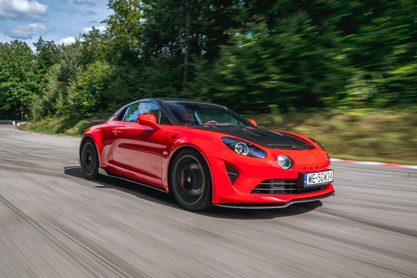 Alpine A110 R to zestrojona z myślą o torowej jeździe odmiana francuskiego modelu Czerwony sportowy samochód z czarnymi detalami jadący dynamicznie po zakręcie toru wyścigowego, otoczony zielonymi drzewami i niebem z lekkimi chmurami.