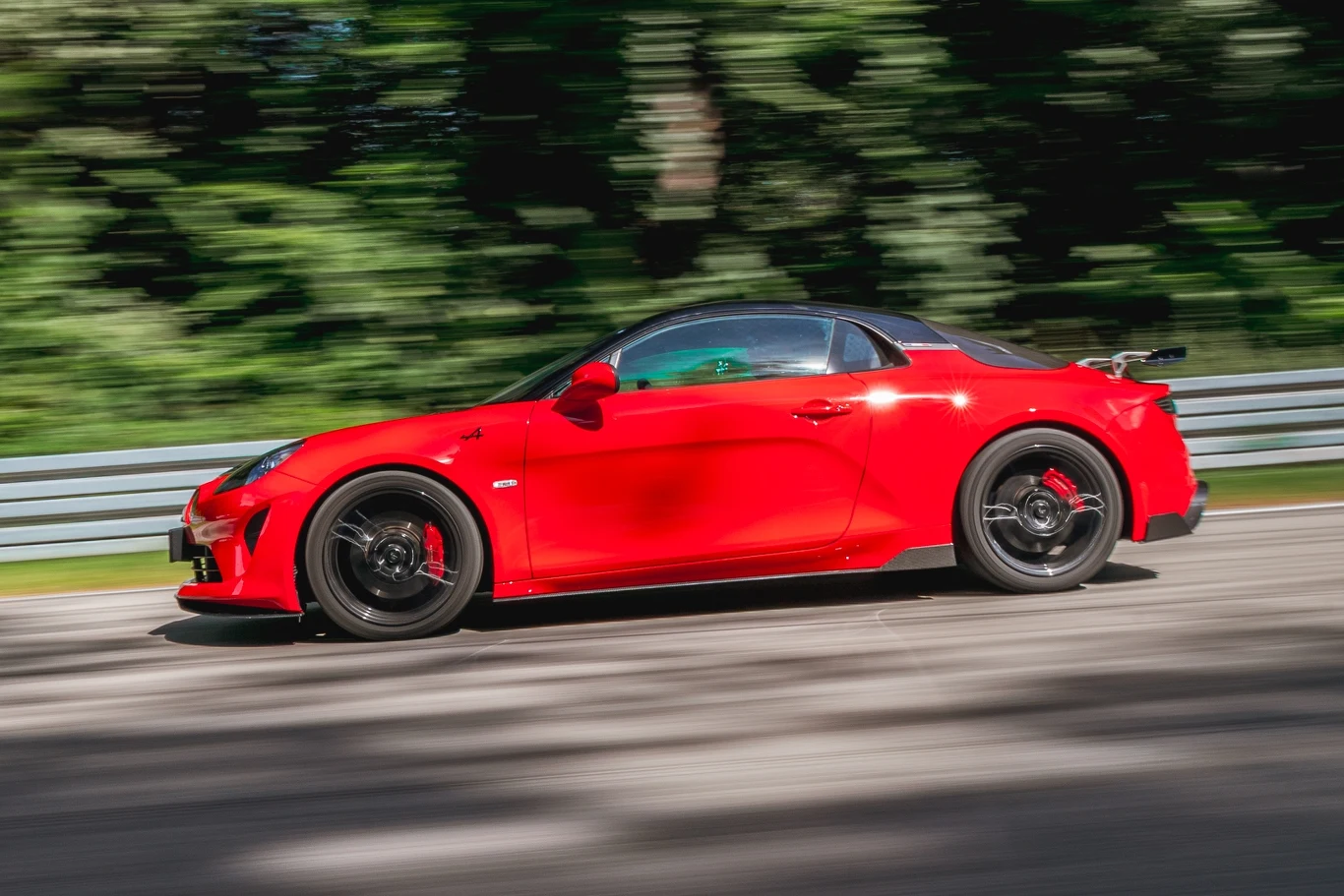 A110 nadal prezentuje się doskonale i ma sylwetkę prawdziwego sportowca Czerwony sportowy samochód jadący z dużą prędkością po asfaltowym torze, otoczony rozmytym, zielonym tłem drzew i metalową barierką ochronną.