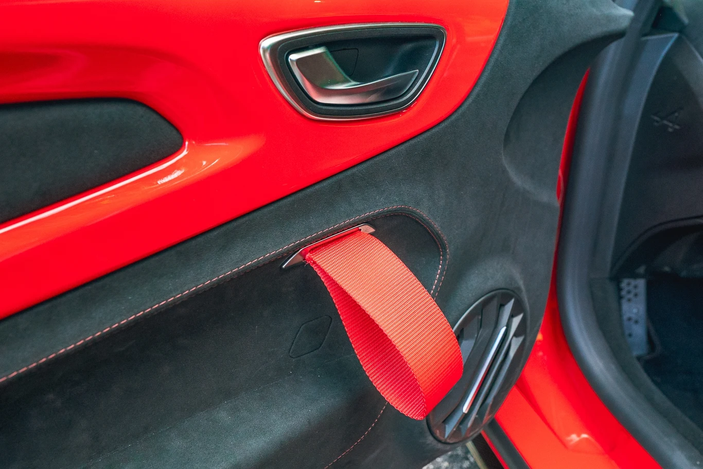 Czerwone taśmy pełnią rolę uchwytów i pozwalają zamknąć drzwi nawet jeśli zapięliśmy już szelki Wnętrze czerwonych drzwi samochodu sportowego z czarnymi wstawkami i widocznym czerwonym paskiem do zamykania drzwi, chromowaną klamką oraz głośnikiem umieszczonym w dolnej części panelu.
