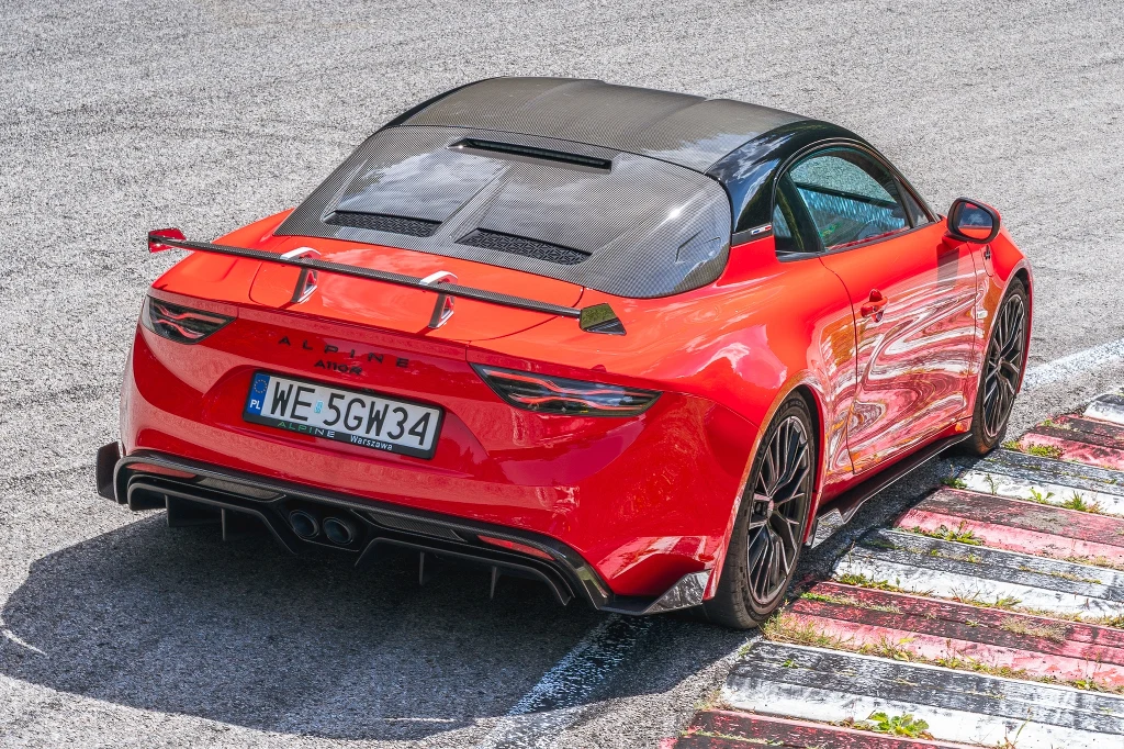 Czerwony sportowy samochód Alpine A110 S na torze wyścigowym, widok z tyłu, samochód stoi przy krawężniku toru, podkreślone sportowe detale takie jak spoiler, dyfuzor oraz czarne felgi, dach częściowo pokryty czarnym malowaniem.