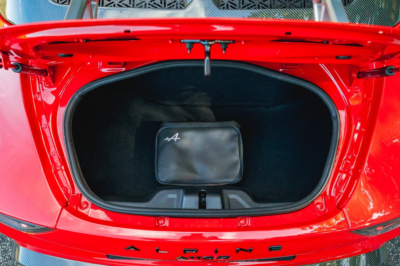 Tylny bagażnik Alpine A110 ma tylko 100 l pojemności, niewielki otwór i nieregularny kształt otwarty bagażnik czerwonego samochodu Alpine A110 z widoczną czarną torbą w środku i logo marki na brzegu bagażnika