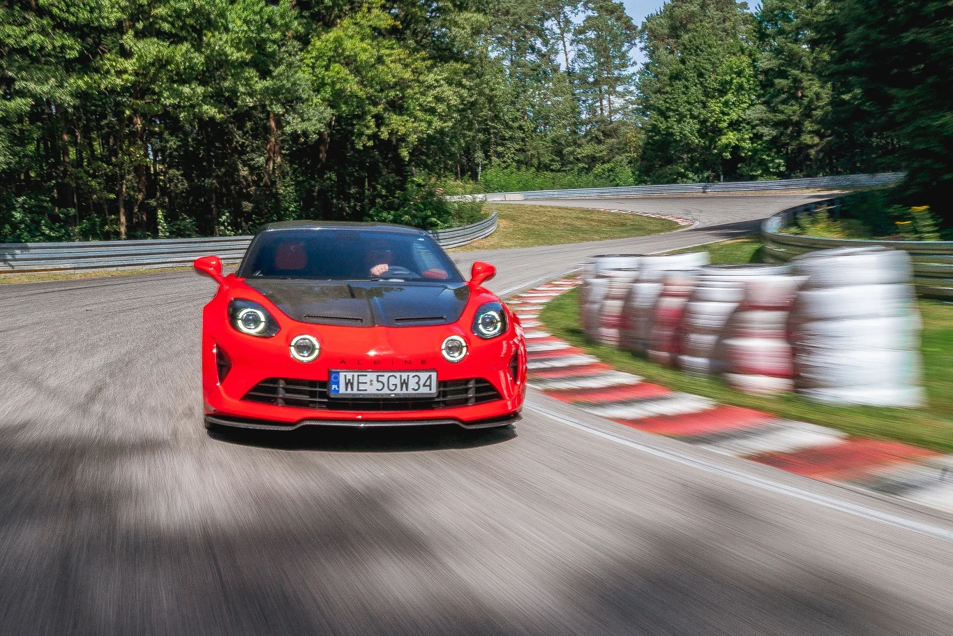 Każde Alpine A110 prowadzi się doskonale, a wersja R ma obniżone o 10 mm zawieszenie, które jest też o 10 proc. bardziej sztywne Czerwony samochód sportowy jadący z dużą prędkością po zakręcie toru wyścigowego, w otoczeniu zieleni lasu i barier ochronnych.