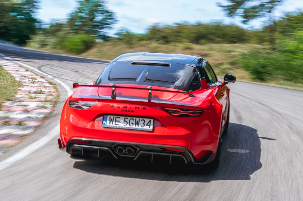 Czerwony sportowy samochód Alpine A110 jadący dynamicznie po zakręcie asfaltowej drogi otoczonej zielenią; dobrze widoczna rejestracja i detale aerodynamiczne pojazdu.