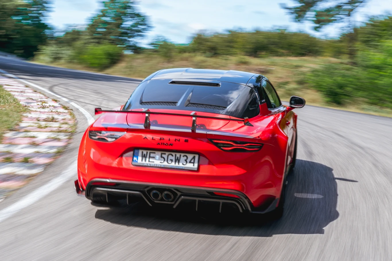 Pakiet aerodynamiczny A110 R zapewnia dodatkowe 29 kg docisku, ale jednocześnie obniżono opory powietrza, co pozwoliło na zwiększenie prędkości maksymalnej do 285 km/h Czerwony sportowy samochód Alpine A110 jadący dynamicznie po zakręcie asfaltowej drogi otoczonej zielenią; dobrze widoczna rejestracja i detale aerodynamiczne pojazdu.