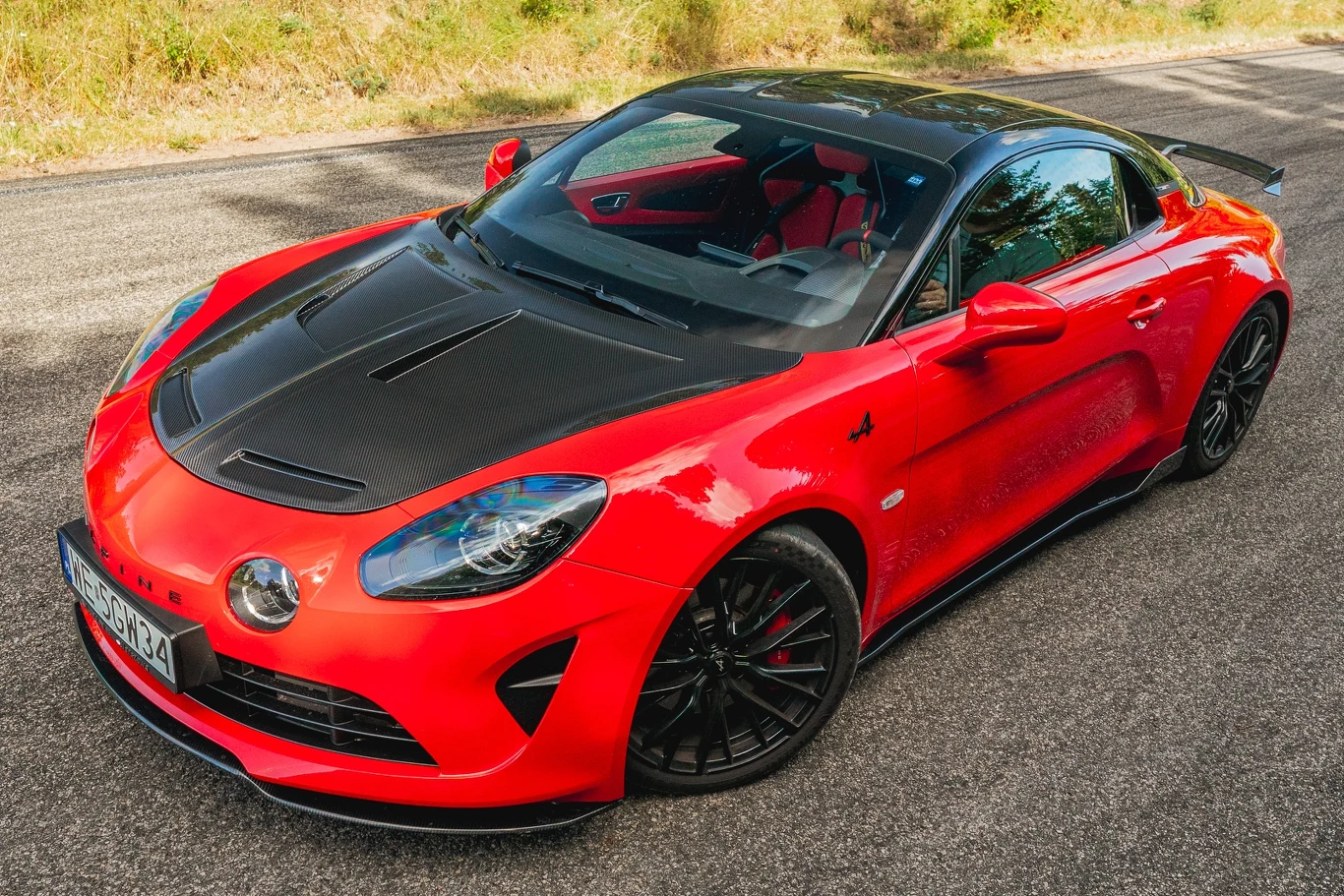 Alpine A110 R prezentuje się doskonale, szczególnie gdy pokryte jest wyrazistym kolorem, kontrastującym z karbonowymi dodatkami Czerwony sportowy samochód z czarnym dachem i maską stoi zaparkowany na asfaltowej drodze, w tle widać fragmenty zieleni i suchej trawy.