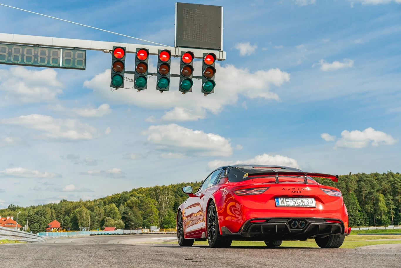 Alpine A110 R najlepiej czuje się na torze Czerwony sportowy samochód stoi na torze wyścigowym pod sygnalizacją świetlną, nad pojazdem widoczny jest zestaw czerwonych świateł charakterystycznych dla startów wyścigowych, w tle las i błękitne niebo z lekkim zachmurzeniem.