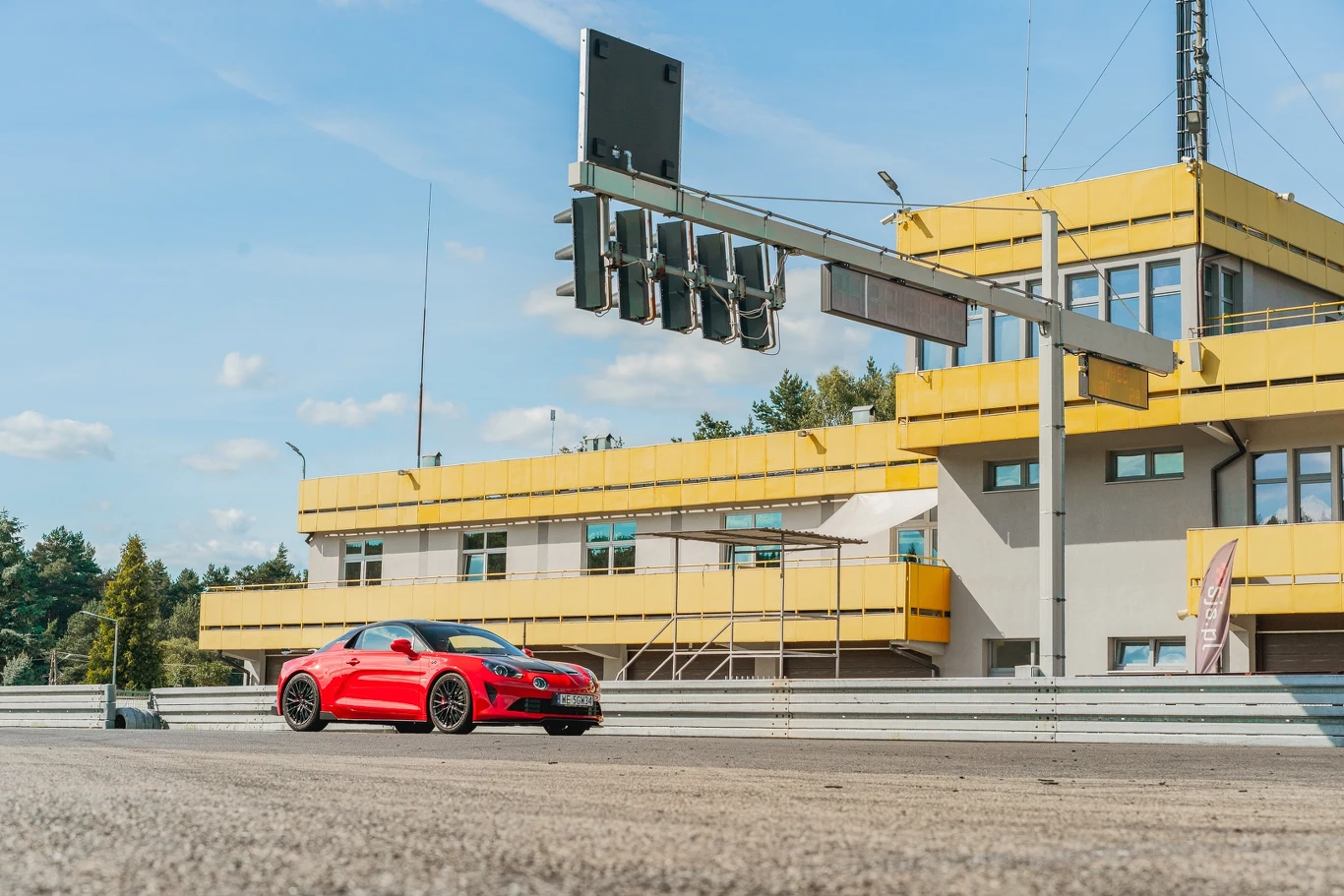 Alpine A110 Czerwony sportowy samochód stoi na torze wyścigowym obok żółtego budynku z trybunami i światłami sygnalizacyjnymi, w tle widoczne drzewa i niebieskie niebo.