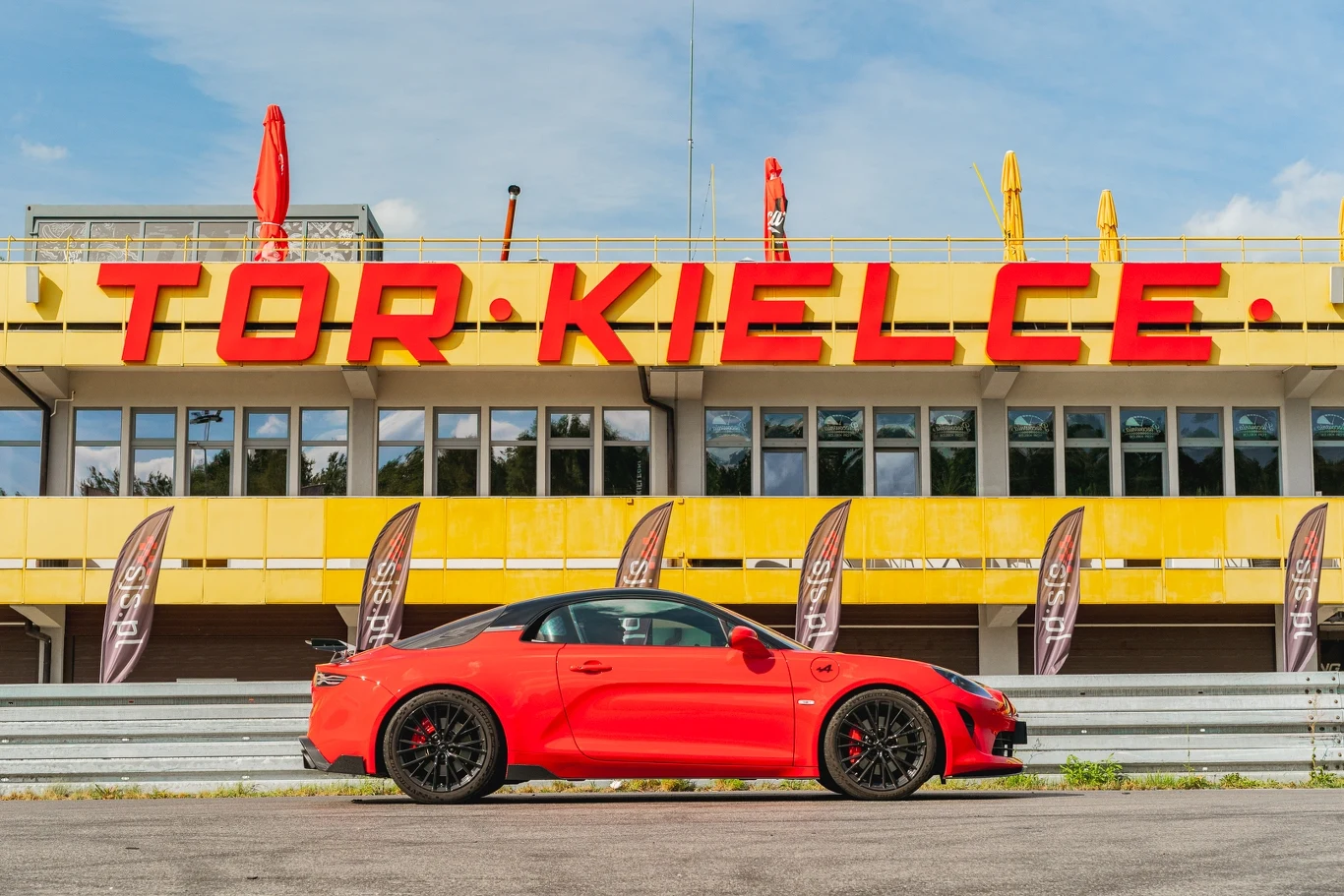 Alpine A110 R Czerwony samochód sportowy zaparkowany przed budynkiem toru wyścigowego Tor Kielce z wyraźnym napisem na żółto-czerwonej elewacji oraz flagami reklamowymi na tle. Słoneczna pogoda i puste trybuny podkreślają motoryzacyjny klimat.