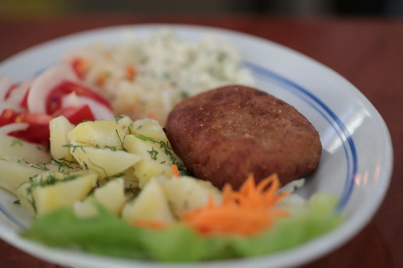 Do dania głównego koniecznie podaj ziemniaki lub kluski śląskie Porcja gotowanych ziemniaków z koperkiem, kotlet mielony, surówka z kapusty oraz plasterki pomidora i cebuli, całość ułożona na białym talerzu.