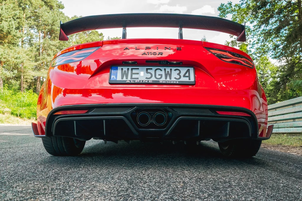 Czerwony samochód sportowy Alpine A110S z dużym tylnym spojlerem stoi na drodze, w otoczeniu zieleni i drzew, widok z tyłu skupia się na układzie wydechowym i rejestracji pojazdu.