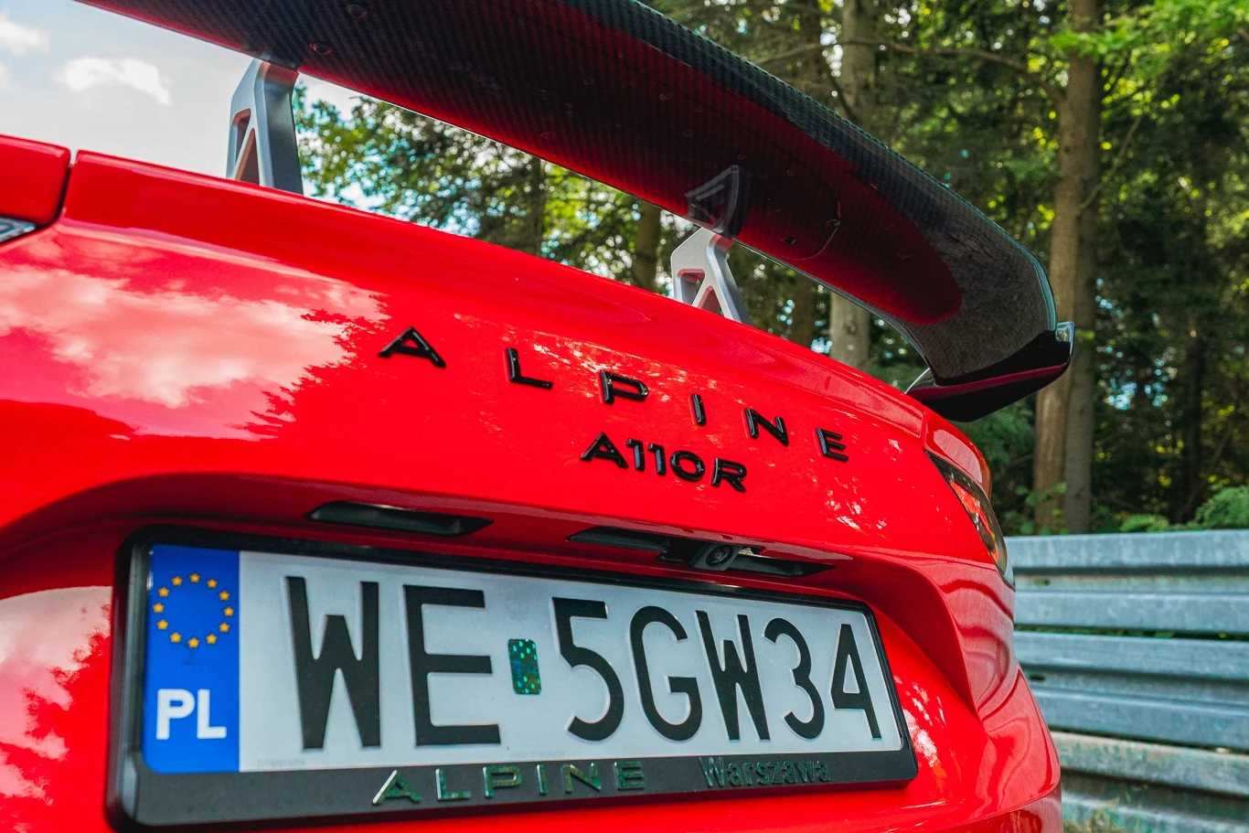 Alpine A110 R Czerwony sportowy samochód marki Alpine A110R z polską tablicą rejestracyjną, widok z tyłu z wyraźnym spojlerem i odbiciem otoczenia w karoserii, w tle drzewa i metalowa bariera ochronna.