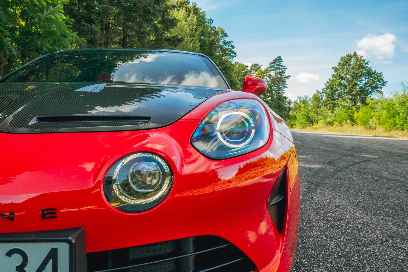 Alpine A110 R Jasnoczerwony sportowy samochód z reflektorami LED ustawiony na asfaltowej drodze, otoczony drzewami i zielenią, z niebieskim niebem i delikatnymi chmurami w tle.