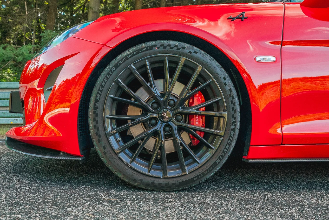 Alpine A110 R Czerwony sportowy samochód z widocznym przednim kołem o czarnych, wieloramiennych felgach na tle asfaltowej drogi i zieleni.