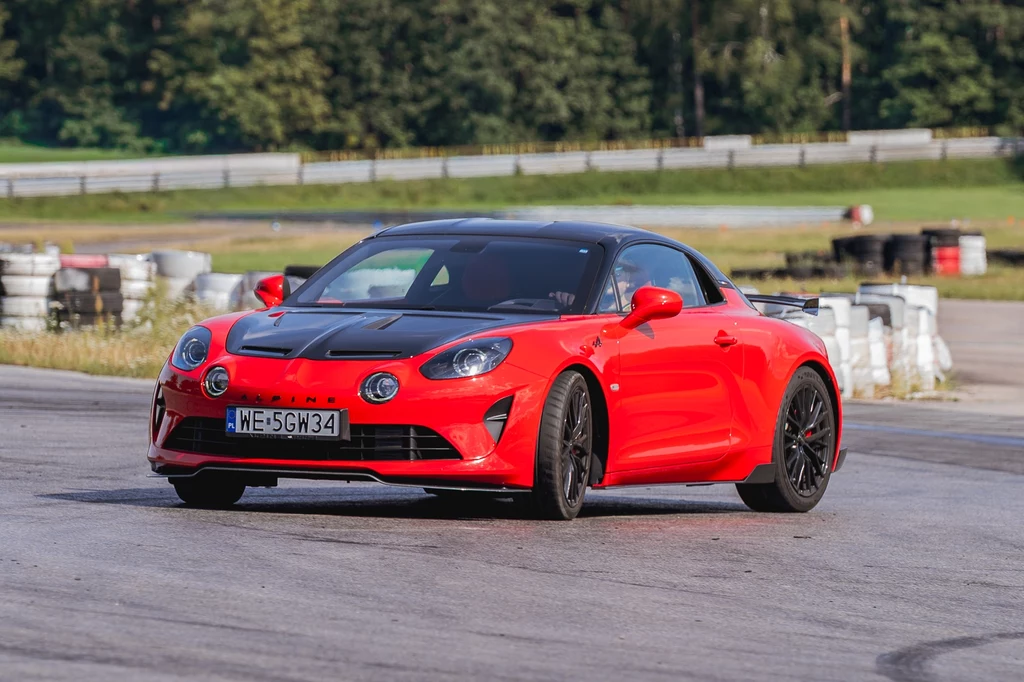 Czerwony sportowy samochód Alpine A110 z czarnymi elementami karoserii pokonuje zakręt na torze wyścigowym, w tle widoczne są bariery z opon oraz zieleń lasu.