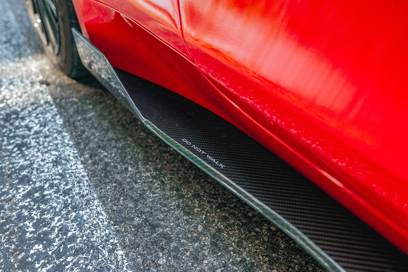 Alpine A110 R Czerwony samochód z widocznym progiem bocznym wykonanym z włókna węglowego, na którym znajduje się napis 'DO NOT WALK', zaparkowany na szaro-białej nawierzchni asfaltowej.