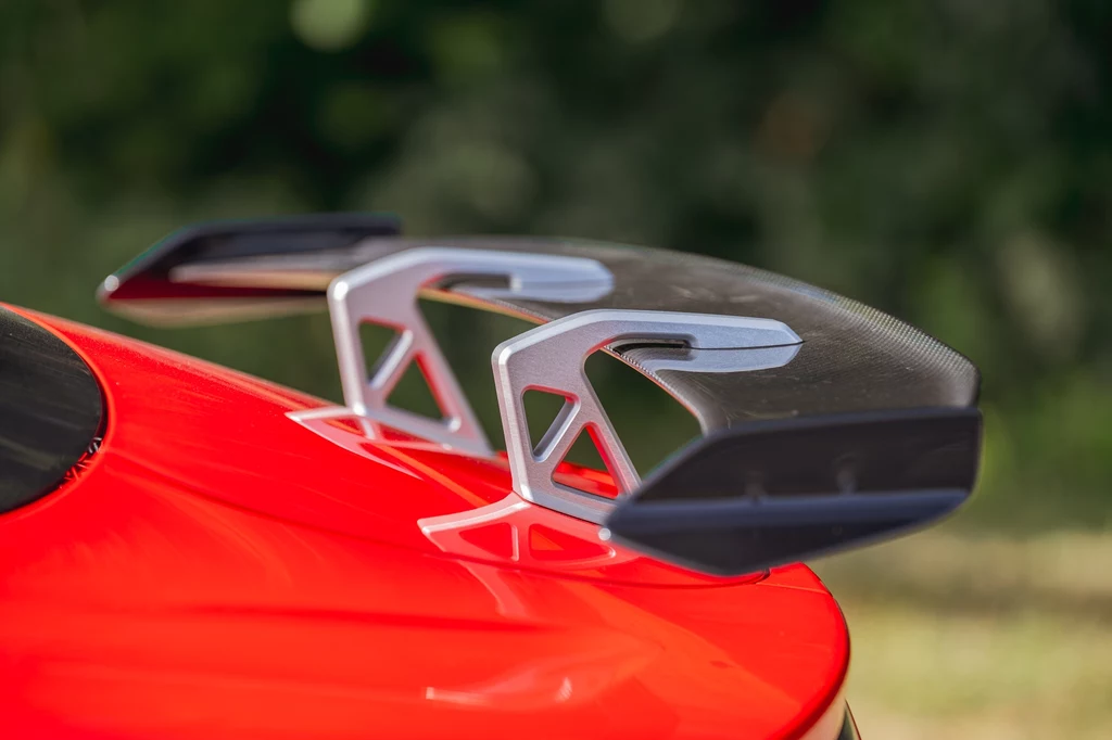 Skrzydło aerodynamiczne wykonane z włókna węglowego zamocowane na tylnej części czerwonego samochodu sportowego z wyraźnie widocznymi wspornikami aluminiowymi.