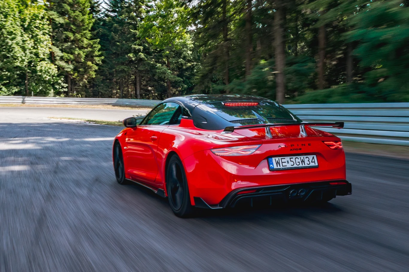 Alpine A110 R Czerwony sportowy samochód jadący z dużą prędkością po asfaltowym torze, wokół gęste drzewa oraz bariera ochronna, samochód wyróżnia się aerodynamicznym kształtem i czarnymi elementami nadwozia.