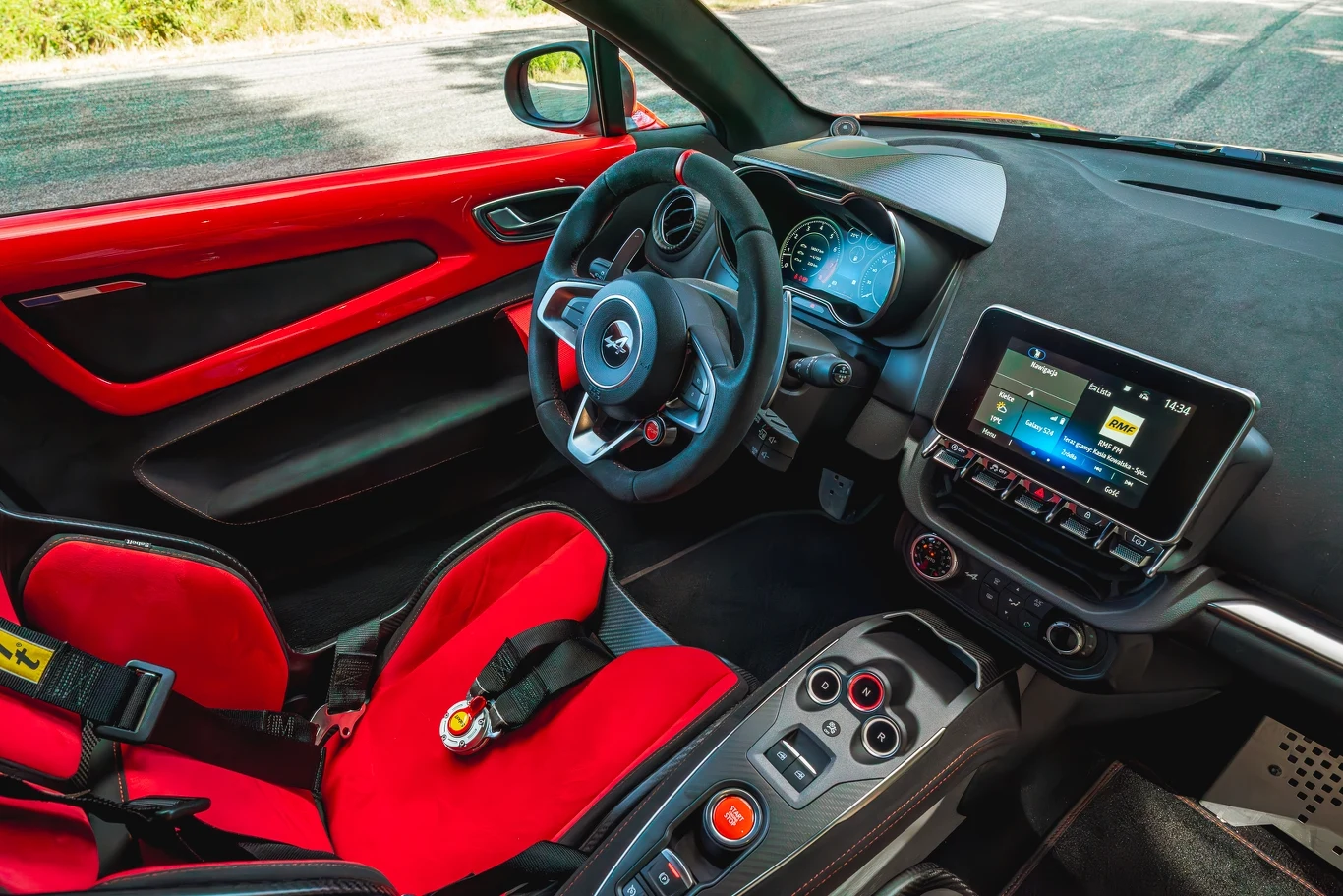 Alpine A110 R Sportowa kabina samochodu z wyprofilowanym czerwonym fotelem kubełkowym, kierownicą z logo i nowoczesną deską rozdzielczą wyposażoną w ekran dotykowy oraz wieloma przyciskami sterującymi.