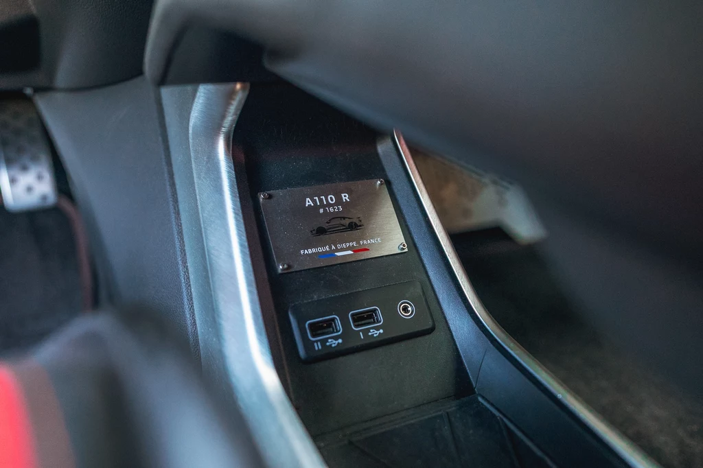 Centralny panel samochodu z widoczną tabliczką znamionową A110 R, dwoma gniazdami USB i detalami wnętrza pojazdu sportowego.
