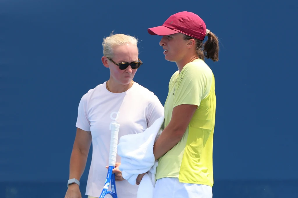 Daria Abramowicz i Iga Świątek Dwie kobiety na korcie tenisowym, jedna ubrana w białą koszulkę i okulary przeciwsłoneczne, druga w żółtej koszulce i czerwonej czapce, trzymający ręcznik i rakietę tenisową, rozmawiają ze sobą na tle niebieskiej ściany.