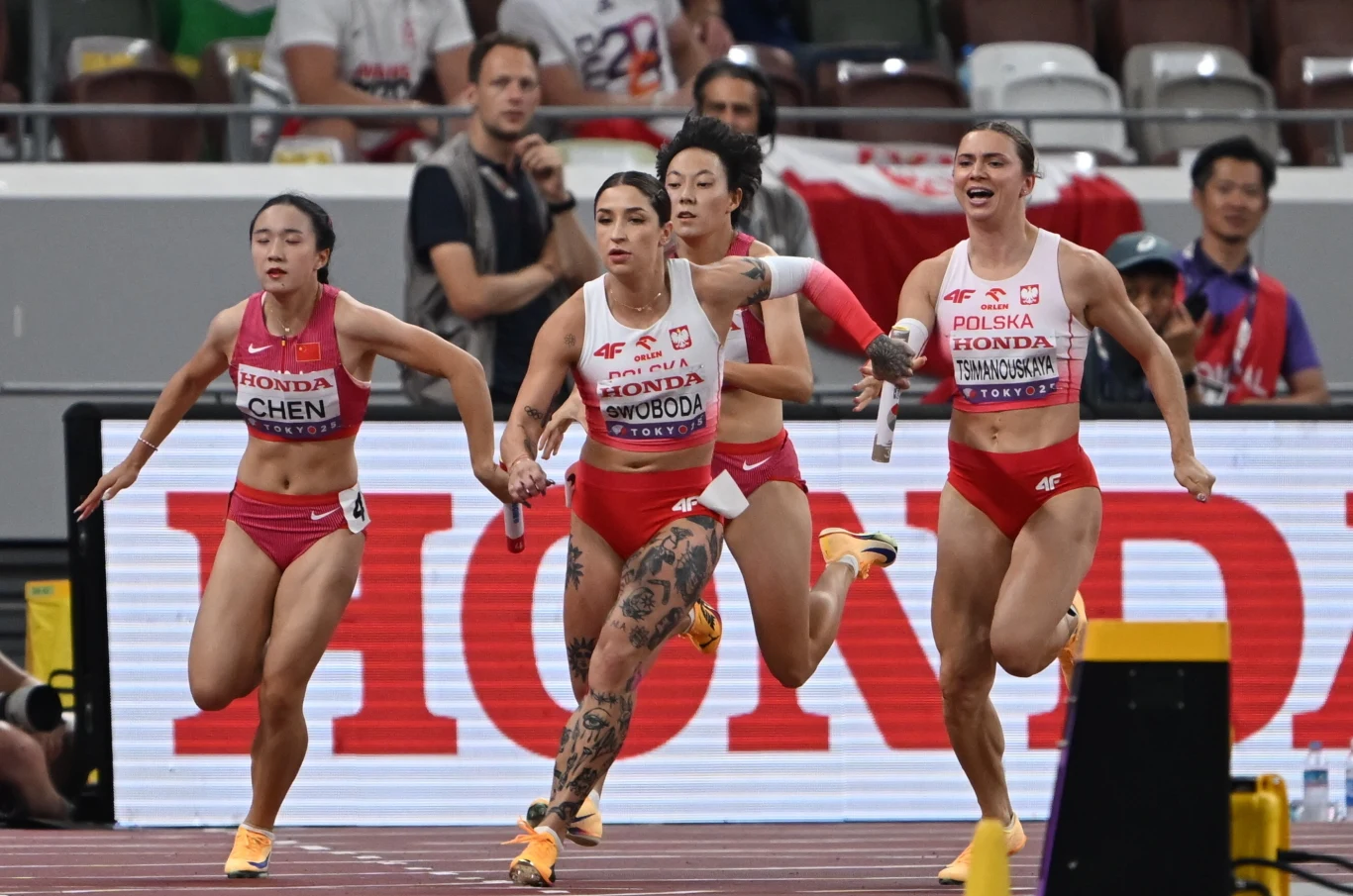 Ewa Swoboda przejmuję pałeczkę od Krysciny Cimanouskowej Zawodniczki biegnące podczas sztafety lekkoatletycznej na stadionie, z widoczną zmianą pałeczki między reprezentantkami Polski i Tajwanu, w tle widzowie oraz duży napis reklamowy.
