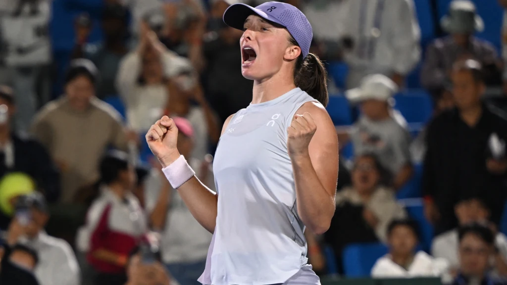 Tenisistka w sportowej czapce i jasnym stroju wyraża silne emocje, triumfując na korcie podczas meczu, w tle widoczna publiczność na trybunach.