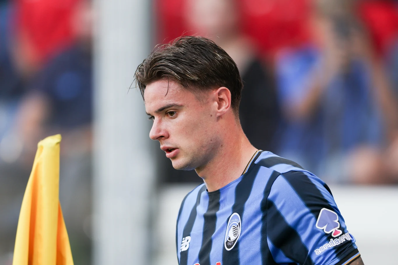 Piłkarz w koszulce w niebiesko-czarne pasy stoi na boisku obok chorągiewki narożnej, w tle rozmyte postacie i czerwone elementy.