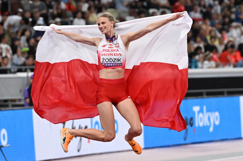Zawodniczka w czerwonych spodenkach i sportowym topie z nazwiskiem Żodzik skacze z szeroko rozpostartą polską flagą, uśmiechając się na tle stadionu lekkoatletycznego wypełnionego kibicami.