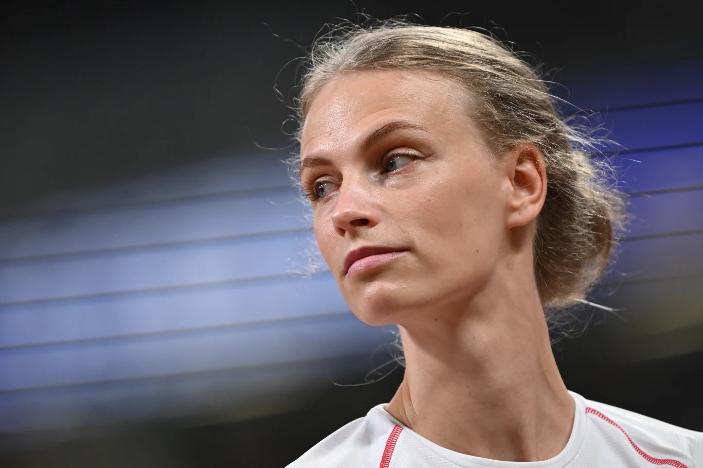 Młoda kobieta z jasnymi włosami spiętymi w kok, ubrana w sportową koszulkę, patrzy w bok z poważnym wyrazem twarzy, w tle rozmyte światła stadionu.