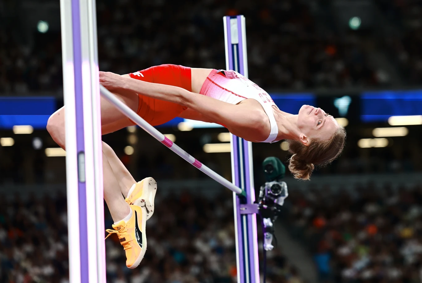 Lekkoatletka w trakcie skoku wzwyż w pełnym napięciu, unosząca się nad poprzeczką podczas zawodów na stadionie wypełnionym publicznością. Skoncentrowana sylwetka zawodniczki w stroju sportowym, dynamiczny ruch oraz wyraziste kolory stroju i sprzętu spo...