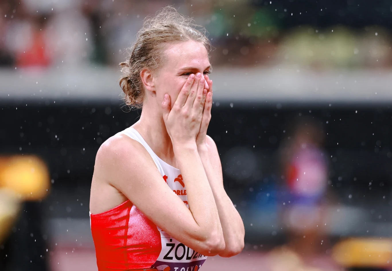 Lekkoatletka w biało-czerwonym stroju sportowym zbliża dłonie do twarzy, okazując silne emocje na tle stadionu w deszczu.