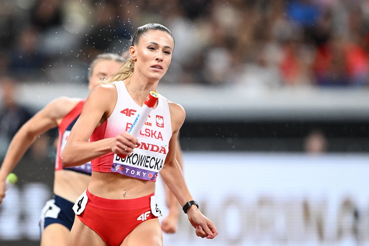 Lekkoatletka w biało-czerwonym stroju reprezentacji Polski biegnie z pałeczką podczas sztafety na stadionie, skupiona na zadaniu, w tle widoczna rywalka.