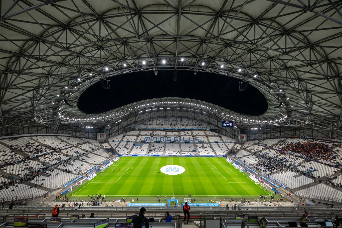 Nowoczesny stadion piłkarski z jasno oświetlonym boiskiem, trybunami częściowo wypełnionymi kibicami oraz dużym logo na środku murawy.