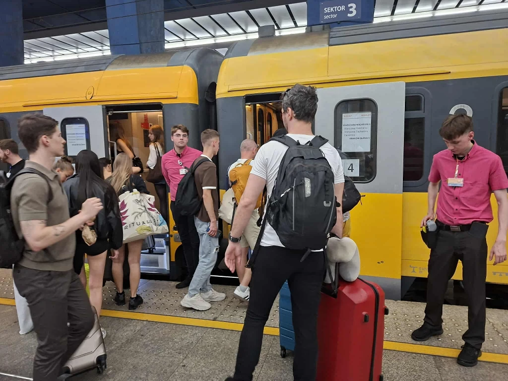 Pasażerowie wchodzący do żółtego pociągu stojącego na peronie, osoby z bagażami, część ludzi w różowych koszulach służbowych, na górze tablica informacyjna z sektorem 3.